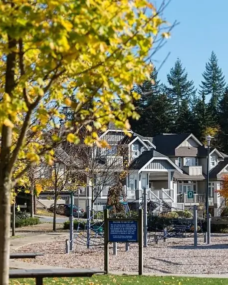 A modern craftsman-style house with dark siding, large windows, a stone foundation, and a landscaped front yard with shrubs and trees. The home features a double garage and is surrounded by autumn foliage.