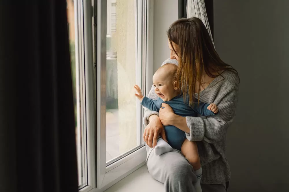 Mother holding child looking out white soundproof replacement window in quiet home